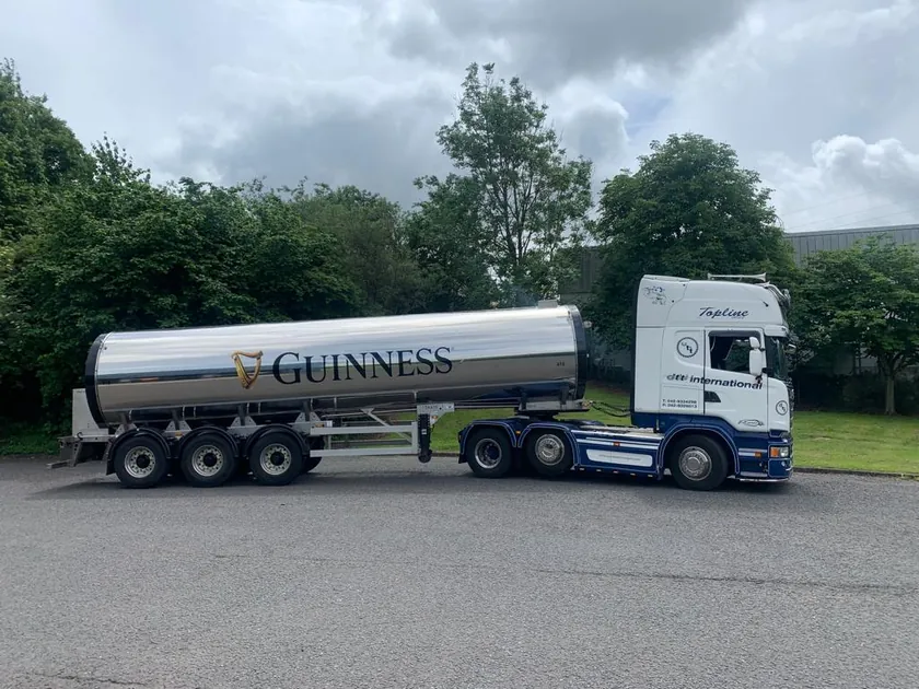 Side view of a polished tanker carrying the Guinness wordmark and harp behind a white and blue truck